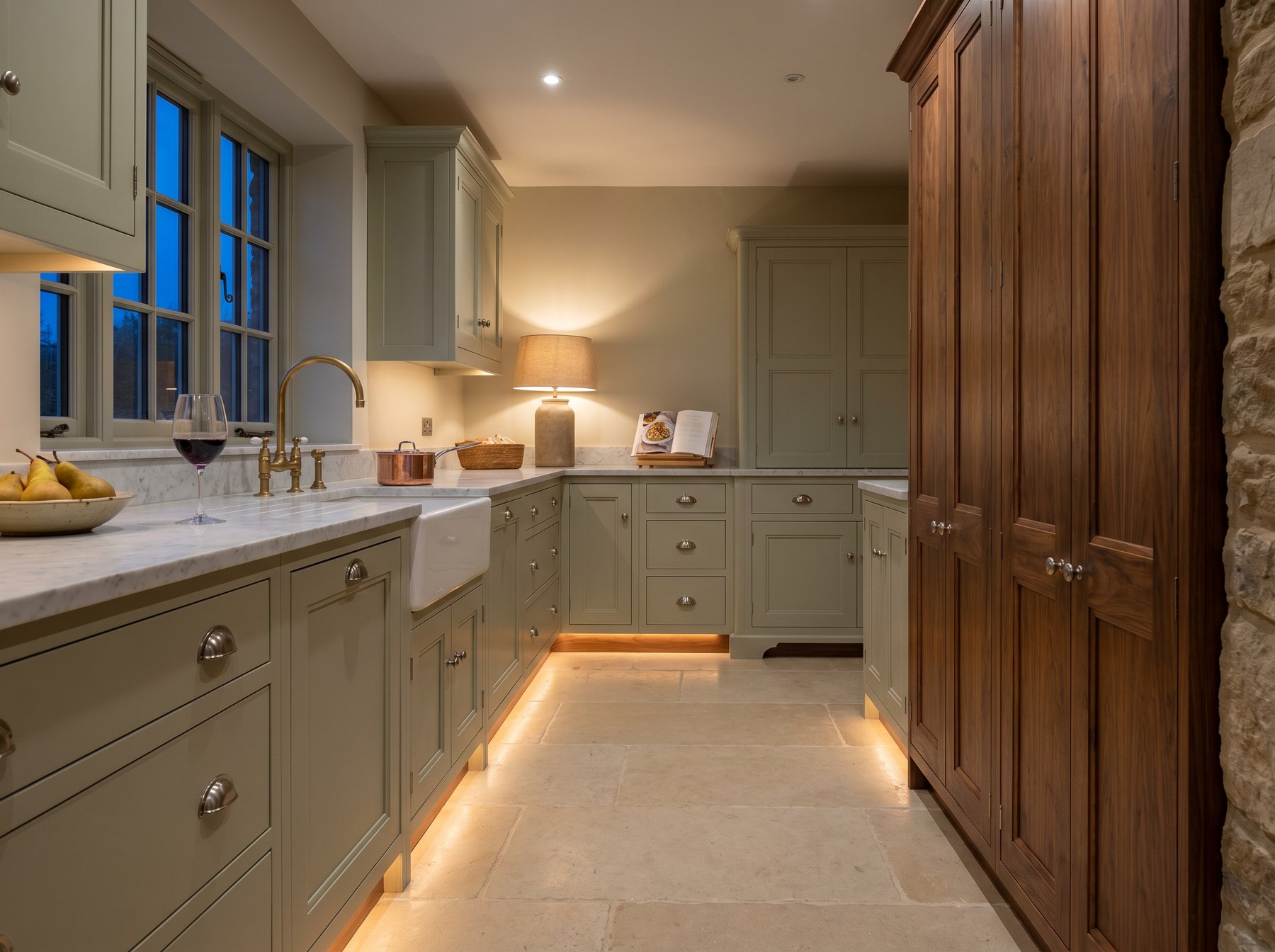 Reeve & Co Suffolk kitchen in soft sage green in-frame Shaker with walnut tall larder, marble worktop and warm plinth lighting at dusk